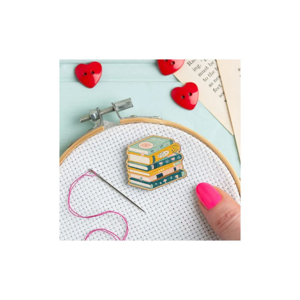 A close-up of a hand with bright pink nails holding an embroidery hoop with white cross-stitch fabric. The fabric features a small embroidered stack of colorful books, surrounded by a needle, pink thread, red heart-shaped buttons, and a piece of paper with printed text.