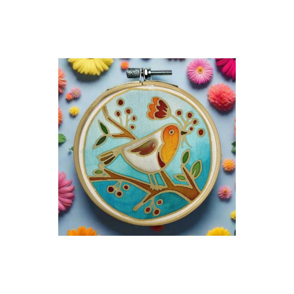 A round embroidery hoop containing a colourful bird design on a light blue fabric background, surrounded by decorative flowers on a blue surface.