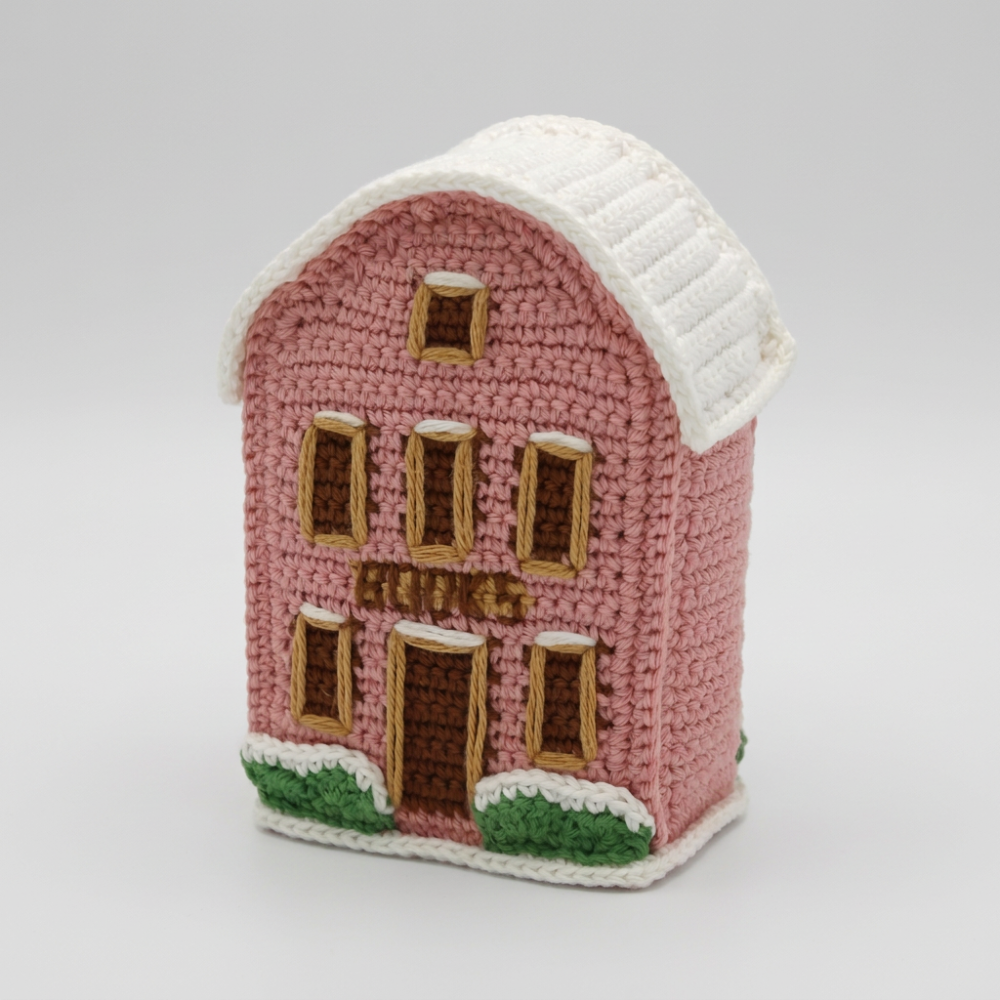 A crocheted house-shaped item, likely a cozy or decorative ornament, in dusty pink with a white roof and trim, brown windows and door, and small green shrubs at the base, shown on a white background.