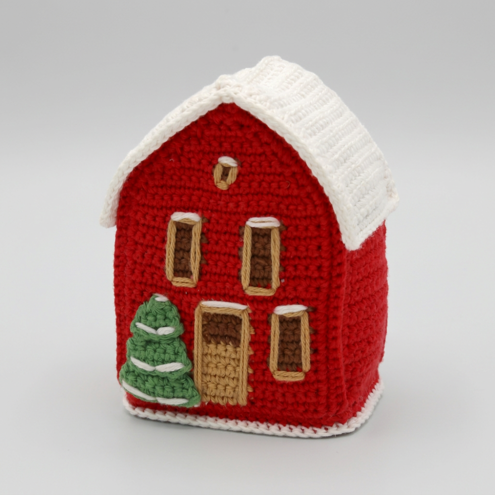 A small crocheted house ornament or decorative figure in red with a white roof, brown window and door details, and a green tree on the front, shown on a plain white background.