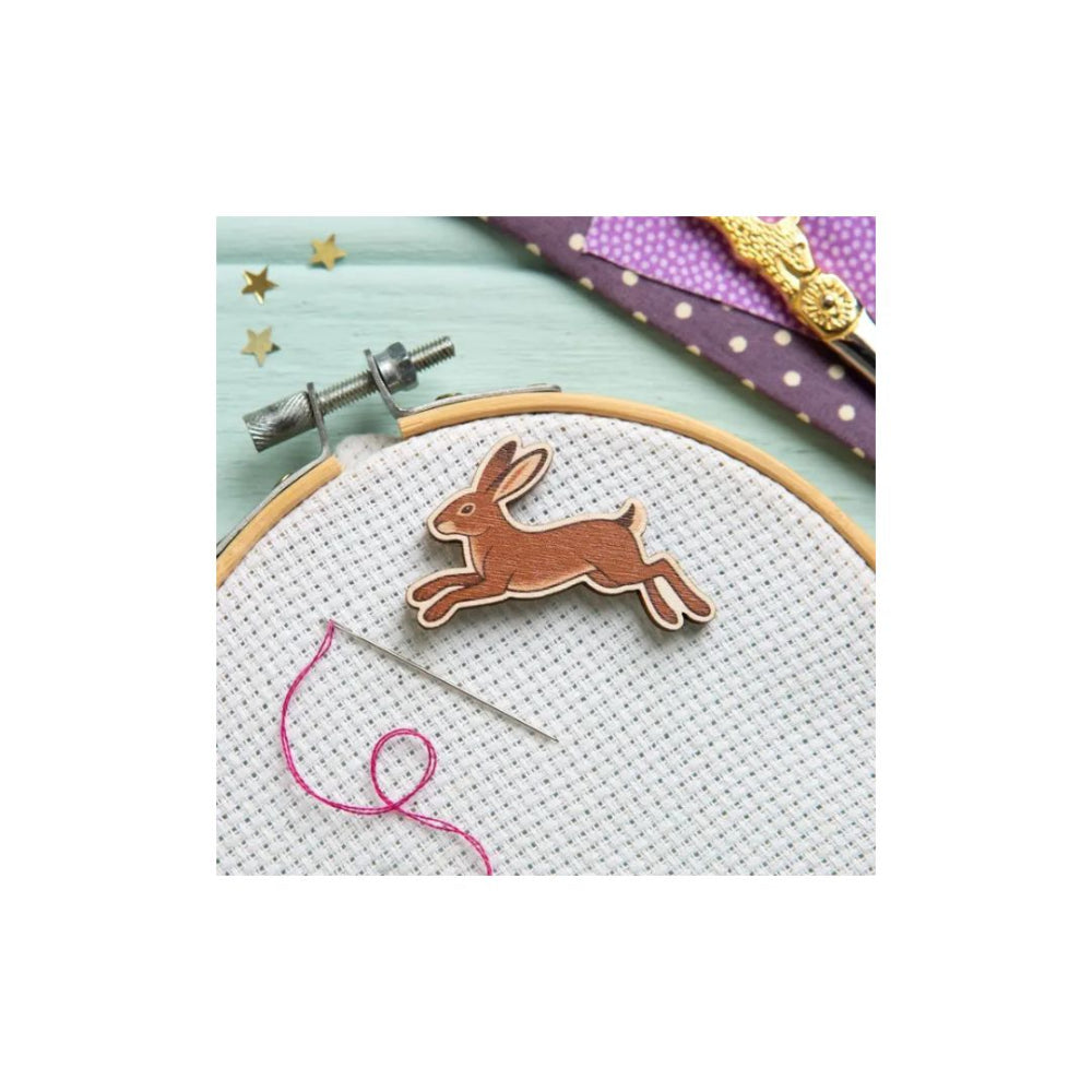 A wooden rabbit-shaped ornament or charm displayed on white cross-stitch fabric inside an embroidery hoop, with a needle, pink thread, and purple patterned ribbon in the background.