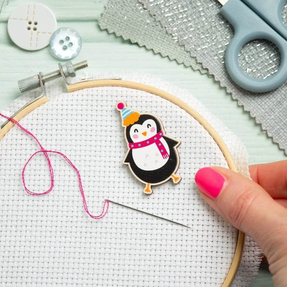 A close-up of a hand with pink nail polish holding an embroidery hoop with white Aida fabric, a wooden needle, pink thread, and a small embroidered penguin patch wearing a party hat and pink scarf. Scissors, buttons, and textured paper are arranged on a pale mint background.