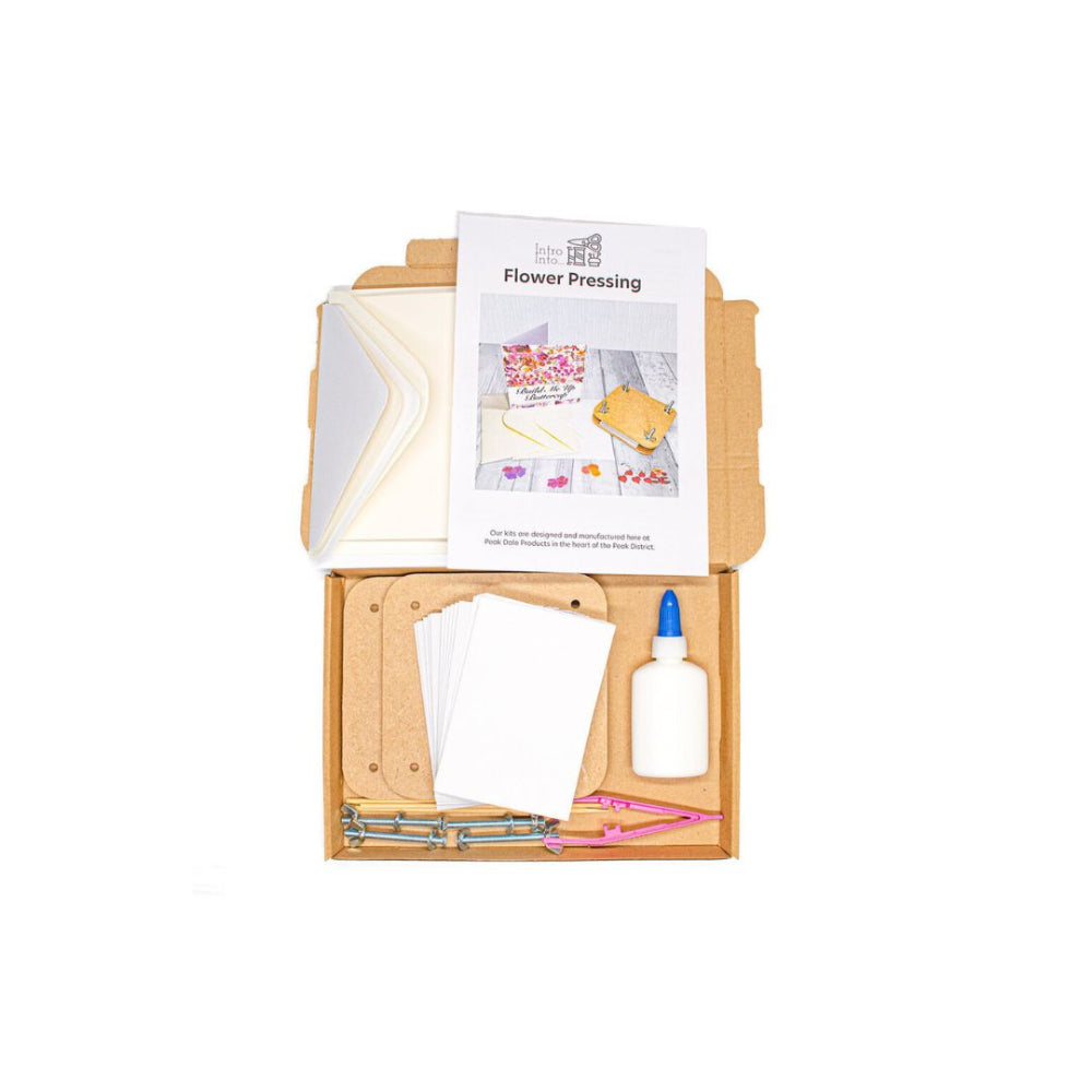 A flower pressing kit displayed inside an open kraft cardboard box on a white background. The kit includes a white envelope, white paper sheets, a small white bottle with a blue cap, metal press bars, pink thread, and a printed instruction sheet titled 'Flower Pressing'.