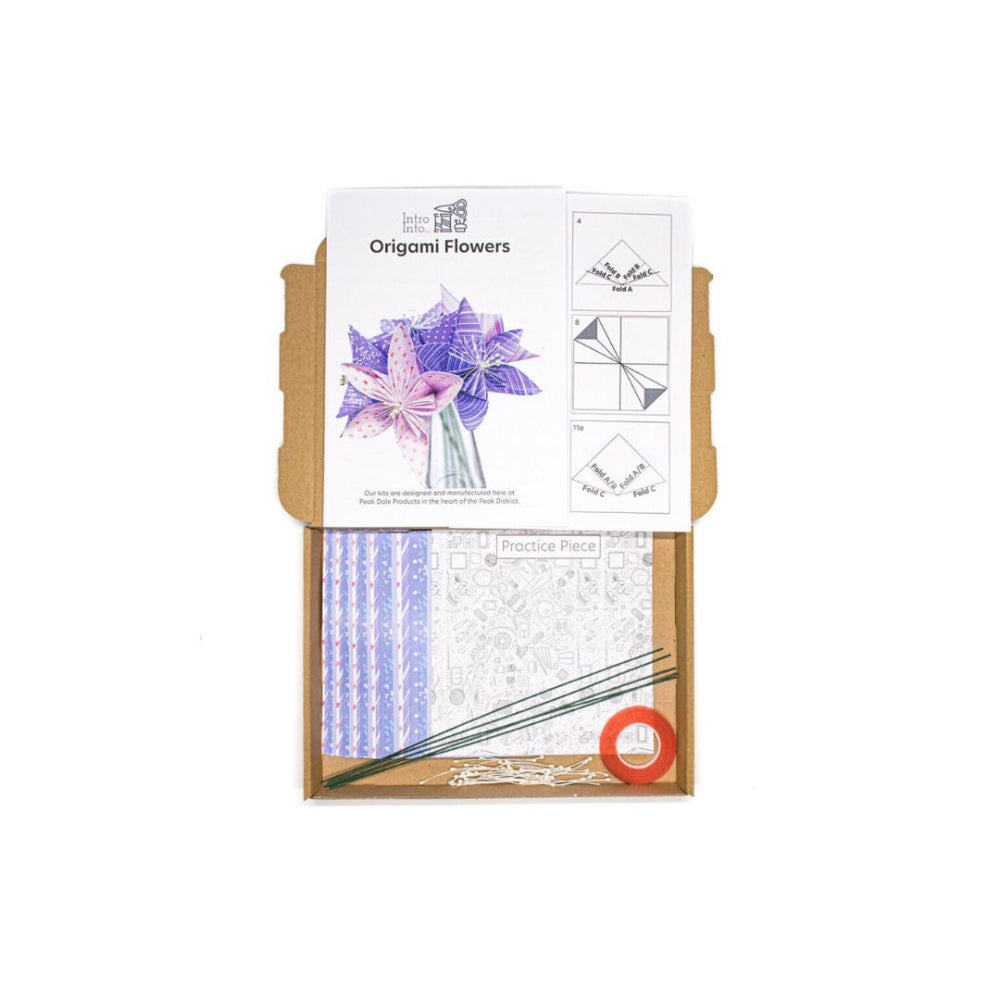 An origami kit in a brown cardboard box, featuring a printed paper template for 'Origami Flowers' with purple and pink lilies, practice sheets, green stems, and a small orange roll, all photographed on a white background.