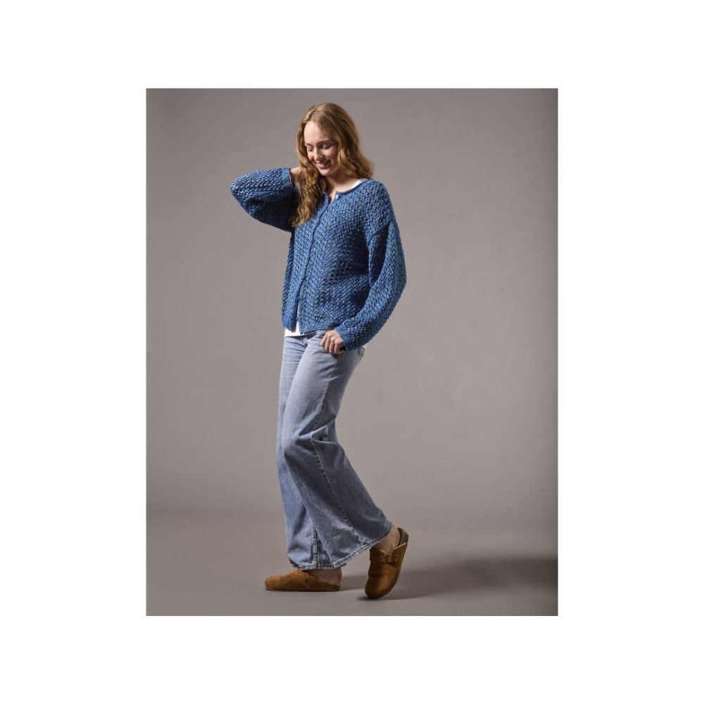 A woman modeling a blue textured knit cardigan sweater, light-wash wide-leg jeans, and brown suede mules against a solid gray studio background.