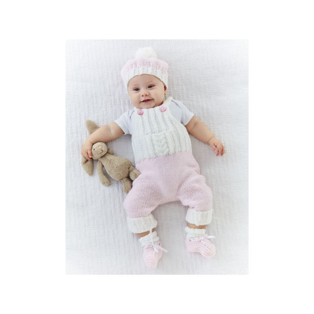 A baby lying on a white textured blanket, wearing a knitted romper, matching hat, and booties, and holding a small stuffed bunny.