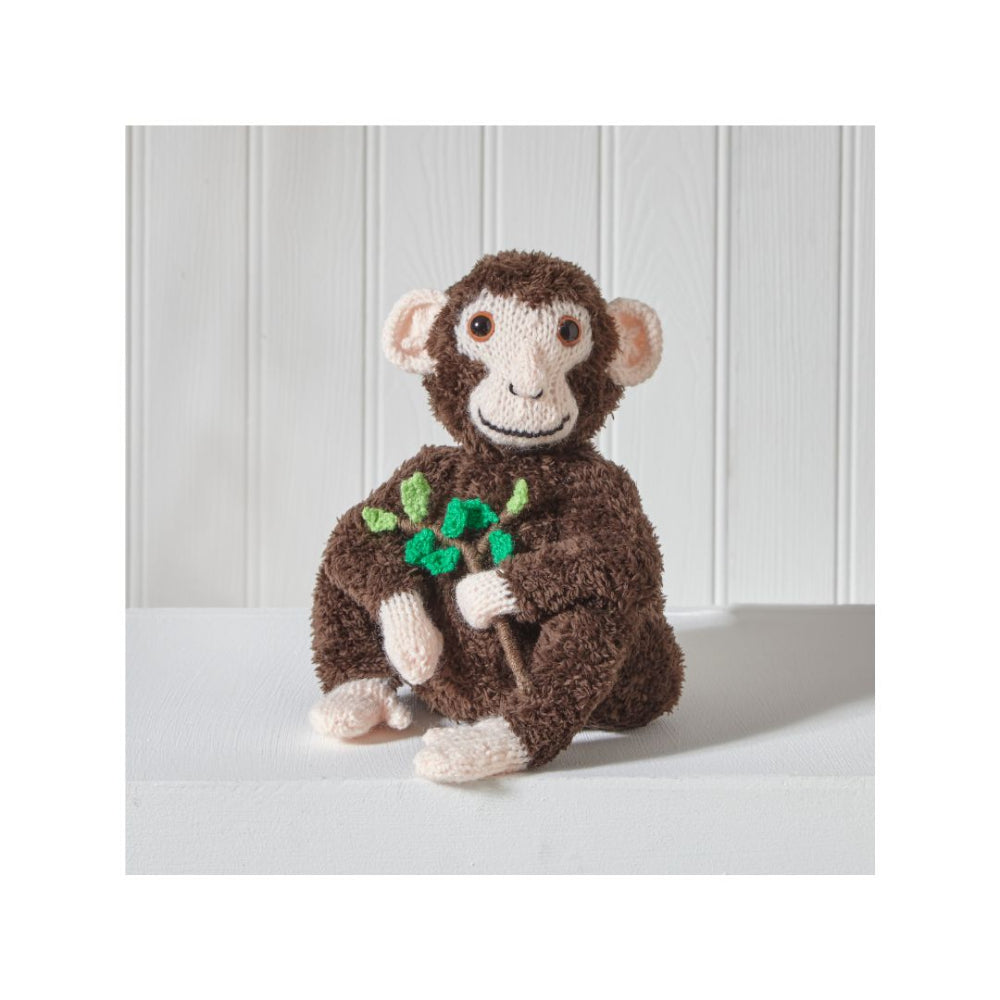 A handmade brown monkey plush toy holding a small green leafy sprig, sitting on a white surface against a white paneled background.