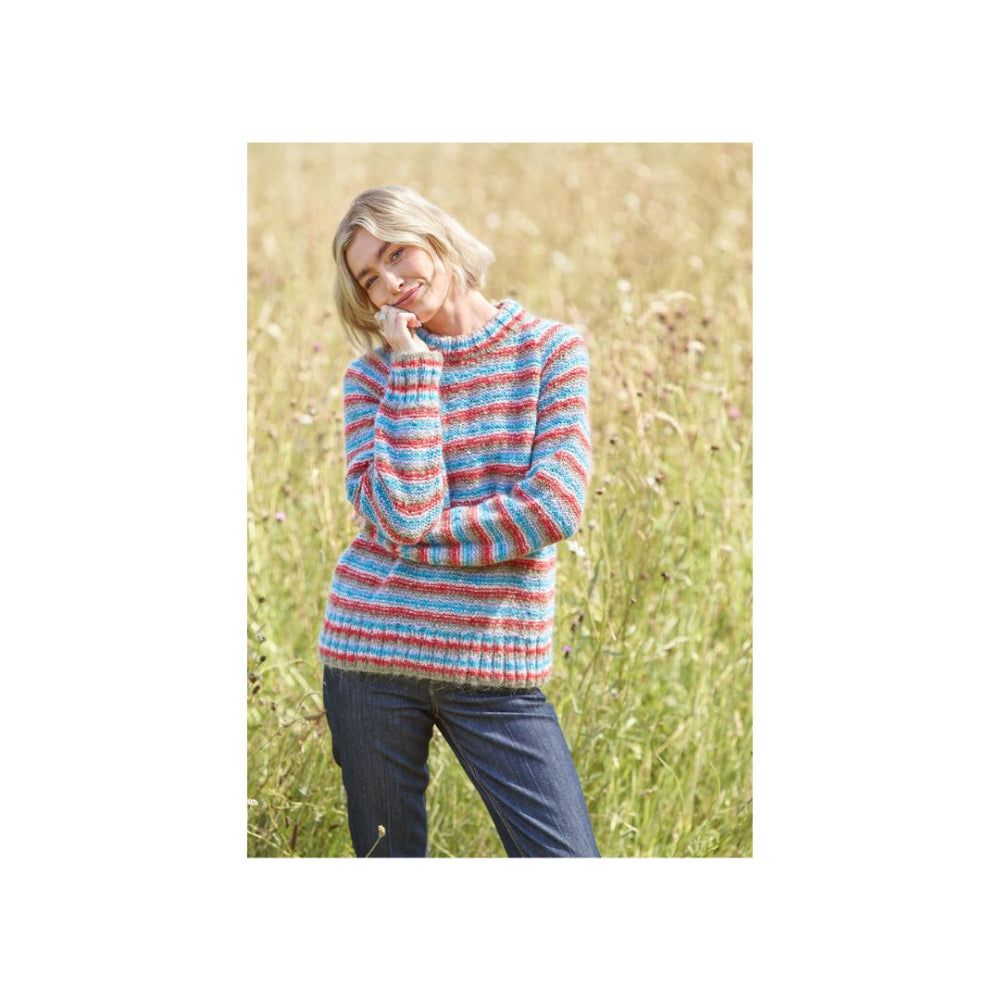 A woman standing in a grassy field wearing a chunky knit sweater with horizontal stripes in blue, pink, and orange, paired with dark blue jeans.