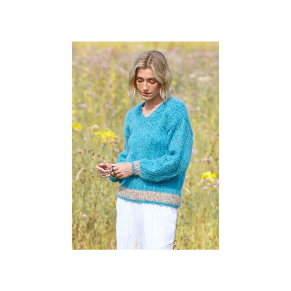 A woman standing in a sunlit field of wildflowers, wearing a fuzzy turquoise blue sweater with a beige ribbed hem and cuffs, paired with white pants.
