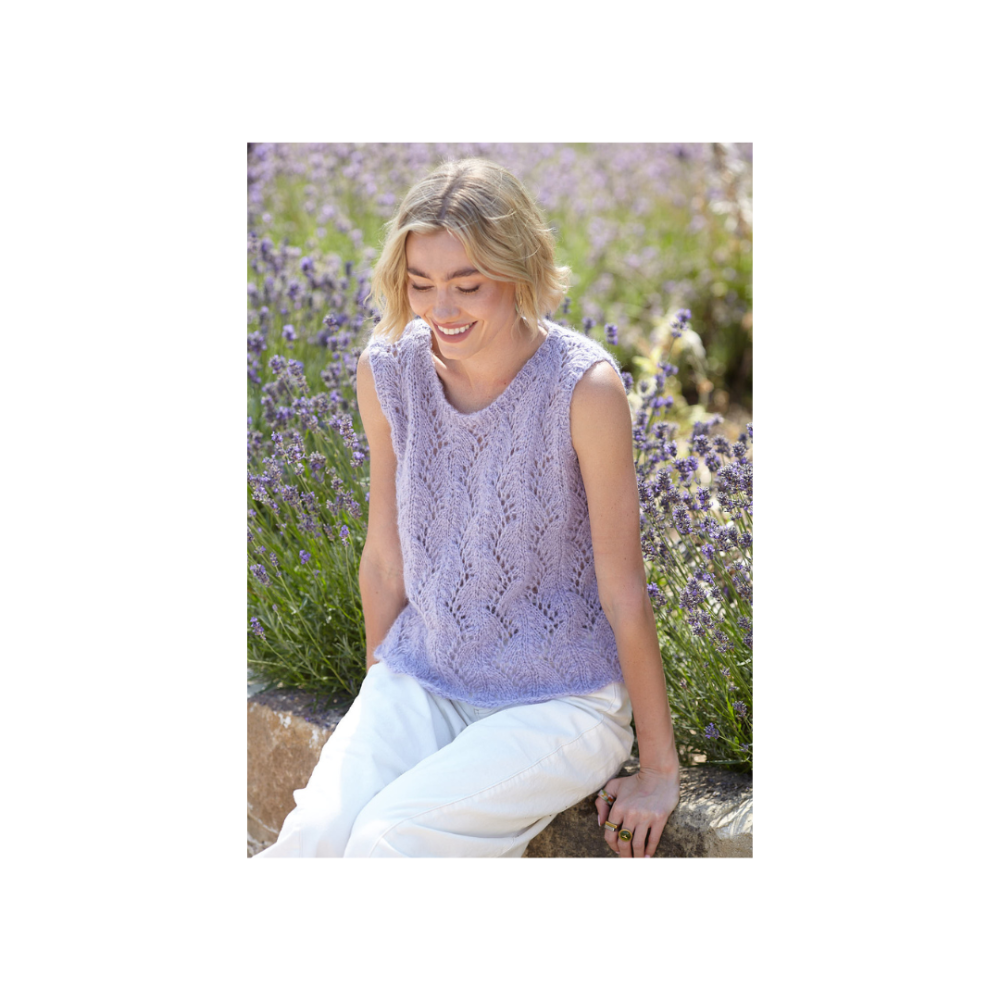 A woman sitting outdoors in a lavender field, wearing a sleeveless light purple knit top and white pants.