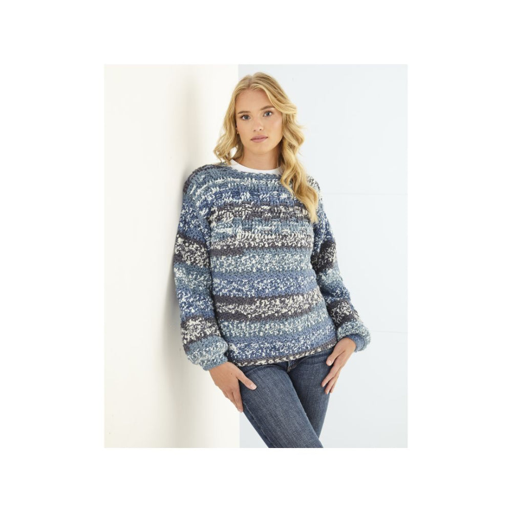 A woman wearing a blue and white marled crochet sweater with horizontal textured stripes, paired with dark blue jeans, leaning against a white wall.
