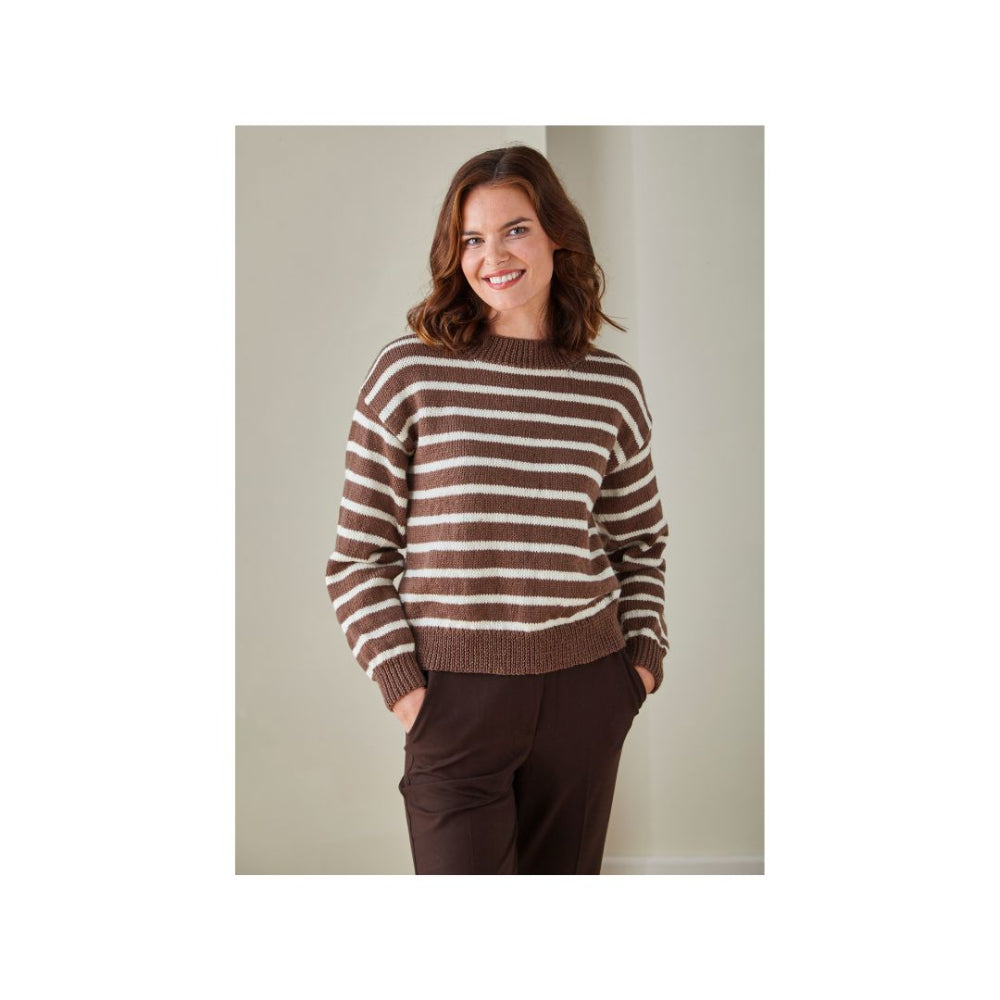 A woman wearing a brown and cream striped knit sweater and dark brown pants, standing against a plain light grey wall.