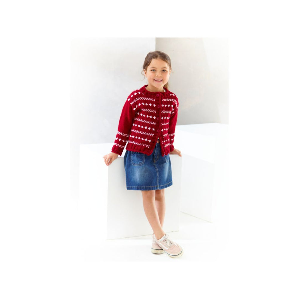 A young girl stands in a bright white studio setting, wearing a red knit cardigan with a white Nordic-style pattern, a blue denim skirt, and light pink sneakers.