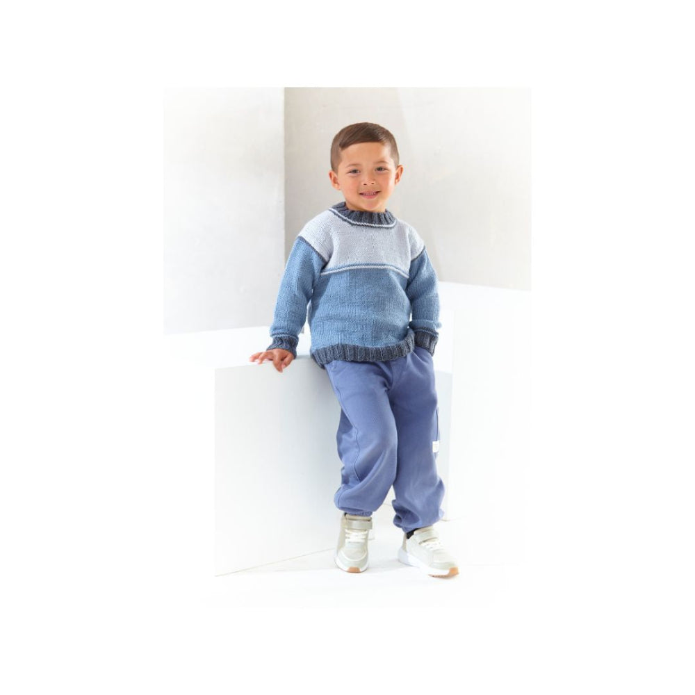A young boy wearing a blue and grey knit round neck sweater and matching blue sweatpants, standing against a white geometric background.