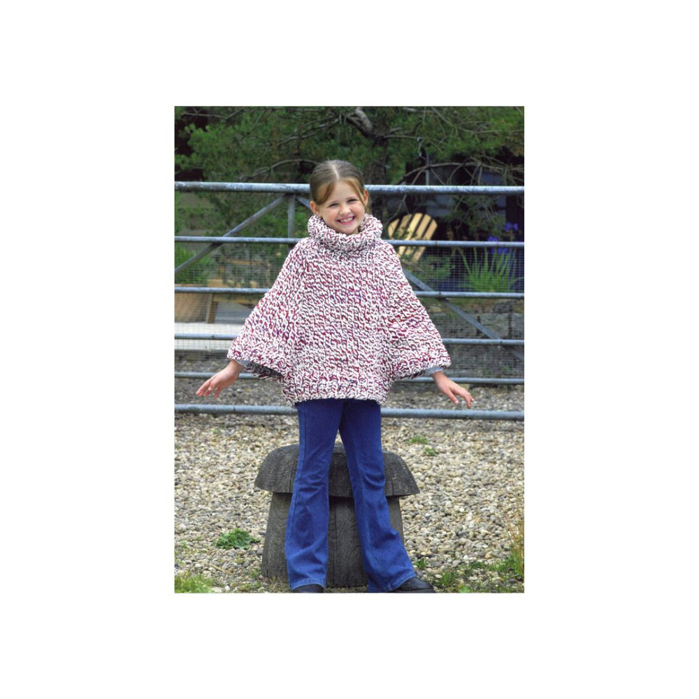 A young girl wearing a chunky knit poncho sweater with a pink, white, and grey pattern, paired with blue pants, standing outdoors on a stone barrel in front of a metal fence.