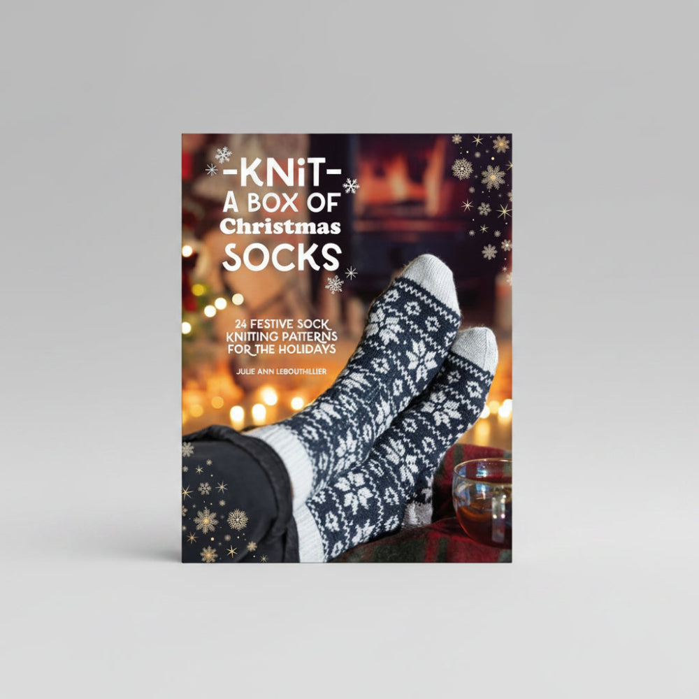 A book or kit cover titled "Knit- A Box of Christmas Socks" showing a pair of knitted Christmas socks on feet against a warm holiday background with text and snowflake graphics.