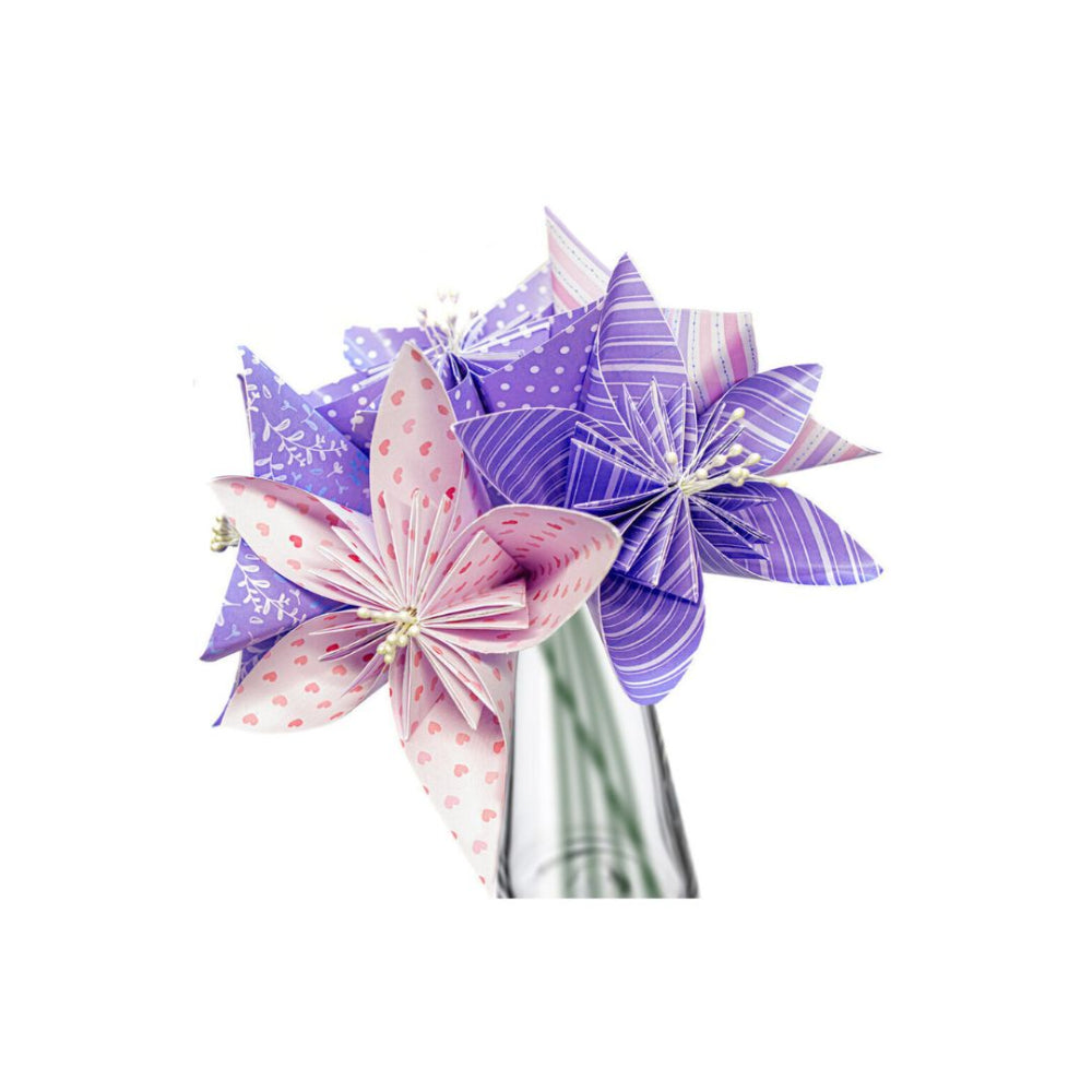 Three handmade origami paper flowers in shades of purple, pink, and white, arranged in a small clear glass vase against a plain white background.