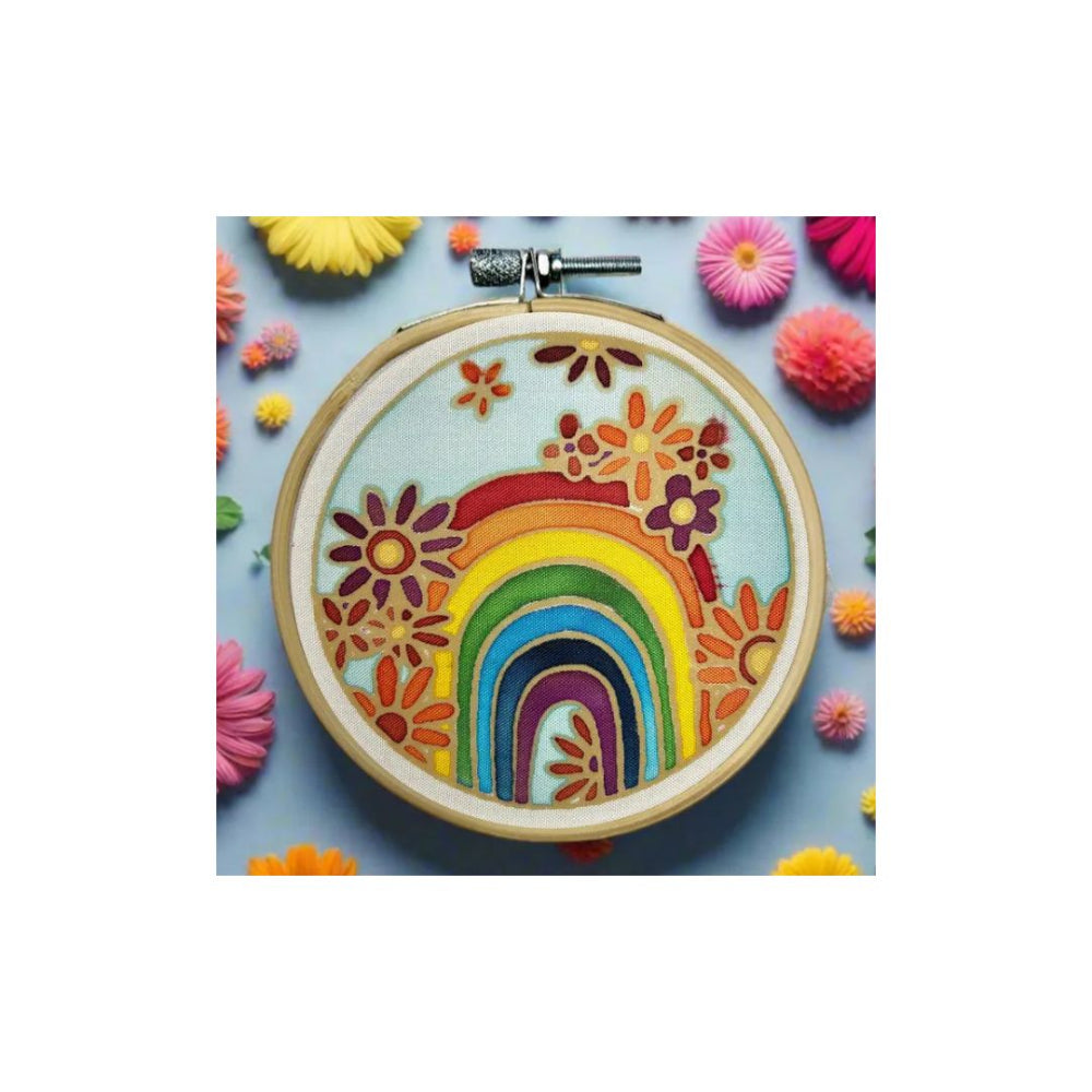 A circular embroidery hoop containing a colourful floral and rainbow design, displayed on a light blue background with decorative paper flowers around it.