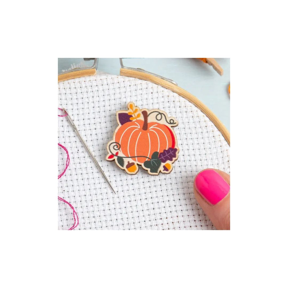 A small embroidered pumpkin charm with autumn leaves and grapes, shown being stitched in a wooden hoop with a needle and a hand with bright pink nail polish.