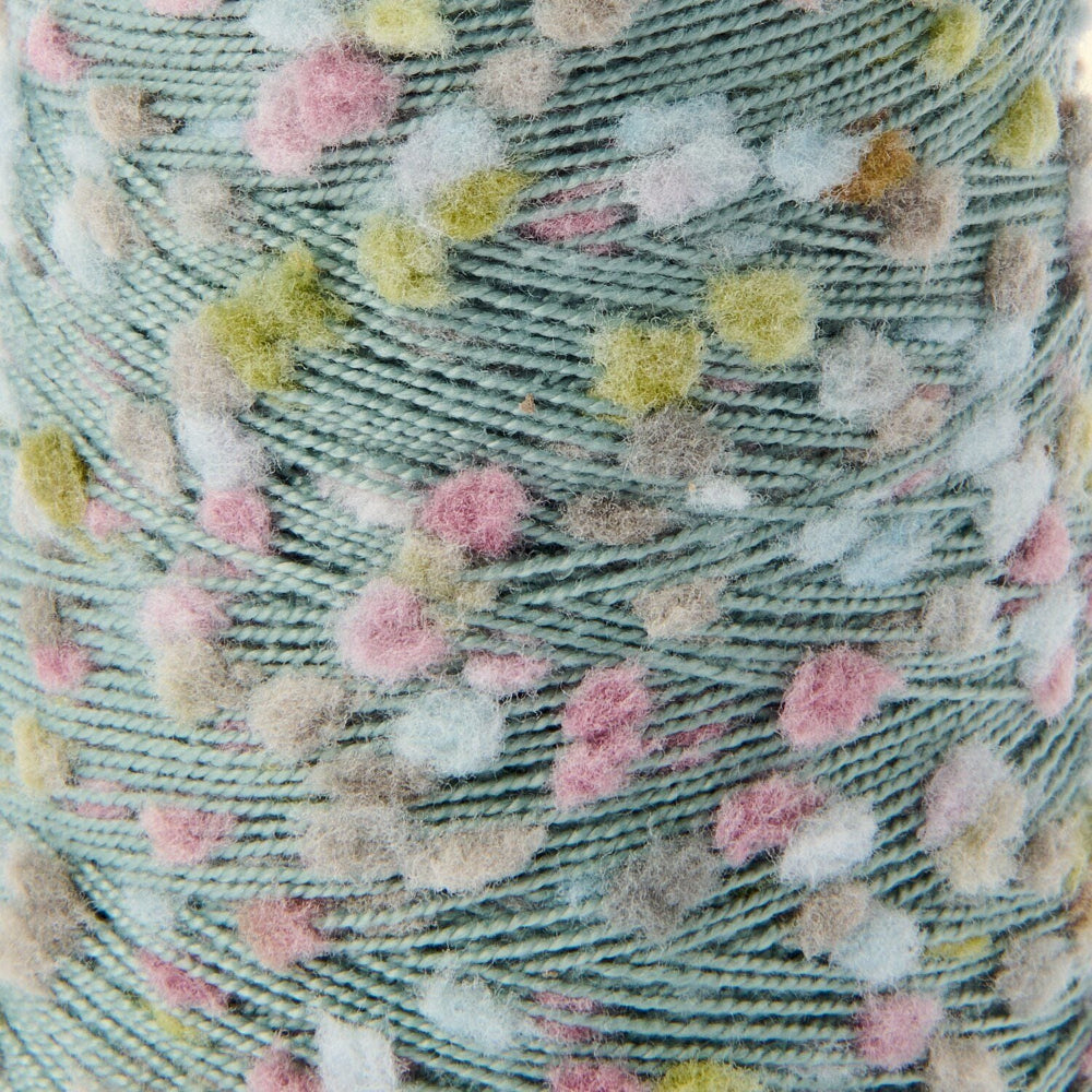 Close-up of a textured yarn spool with a dusty sage green base and scattered fuzzy pom-pom dots in pink, yellow, beige, and white.