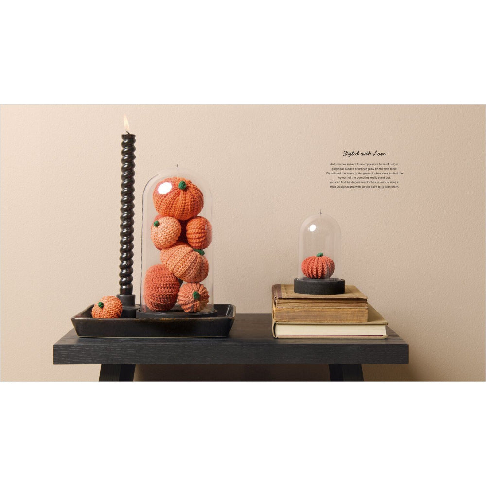 A styled tabletop arrangement with crocheted orange pumpkins under clear domes, a tall black spiral candle, and books against a beige wall. Small text on the wall reads 'Styled with Love'.