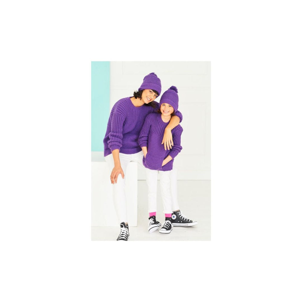 A woman and a child wearing matching purple knit sweaters, beanies, and white pants, standing together against a light studio background.
