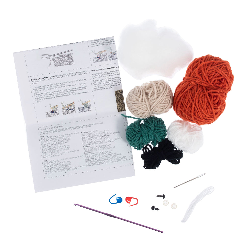 A crochet kit laid out on a white background, including a printed instruction booklet, multiple balls of yarn in orange, cream, green, black, and white, a purple crochet hook, blue and red stitch markers, buttons, a needle, and other small accessories.