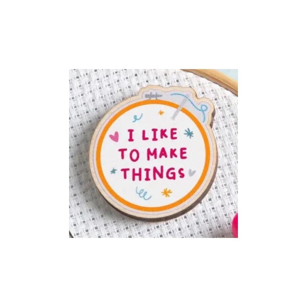 A round wooden embroidery hoop pin with the phrase 'I LIKE TO MAKE THINGS' in pink lettering, surrounded by small pink, blue, and yellow doodles, displayed on white cross-stitch fabric.