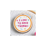 A round wooden embroidery hoop pin with the phrase 'I LIKE TO MAKE THINGS' in pink lettering, surrounded by small pink, blue, and yellow doodles, displayed on white cross-stitch fabric.