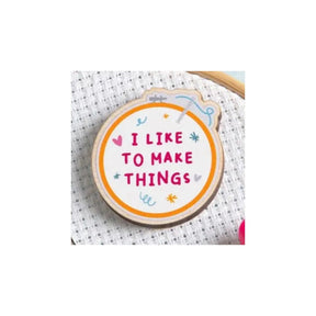 A round wooden embroidery hoop pin with the phrase 'I LIKE TO MAKE THINGS' in pink lettering, surrounded by small pink, blue, and yellow doodles, displayed on white cross-stitch fabric.
