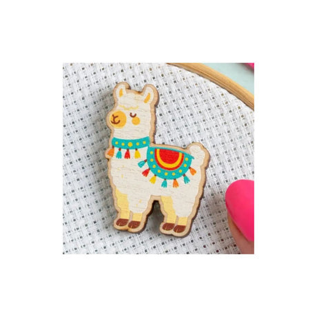 A wooden llama-shaped craft piece with painted details, resting on a white cross-stitch fabric background with a pink nail visible at the edge.