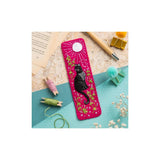 A bright pink embroidered bookmark featuring a black cat surrounded by green leaves and white flowers, with a white sun motif at the top. The bookmark is displayed in a styled flat-lay arrangement on a light blue and beige background with sewing and reading props.