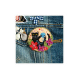 A round embroidered patch featuring a black cat and a blue bird among colourful flowers, shown pinned onto a blue denim jacket.