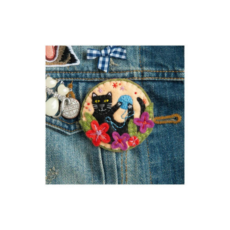 A round embroidered patch featuring a black cat and a blue bird among colourful flowers, shown pinned onto a blue denim jacket.