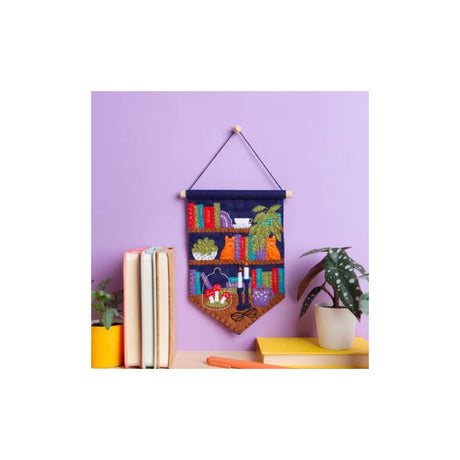 A decorative fabric wall hanging with a colourful bookshelf design, suspended by a string on a lavender wall above a shelf with books and potted plants.