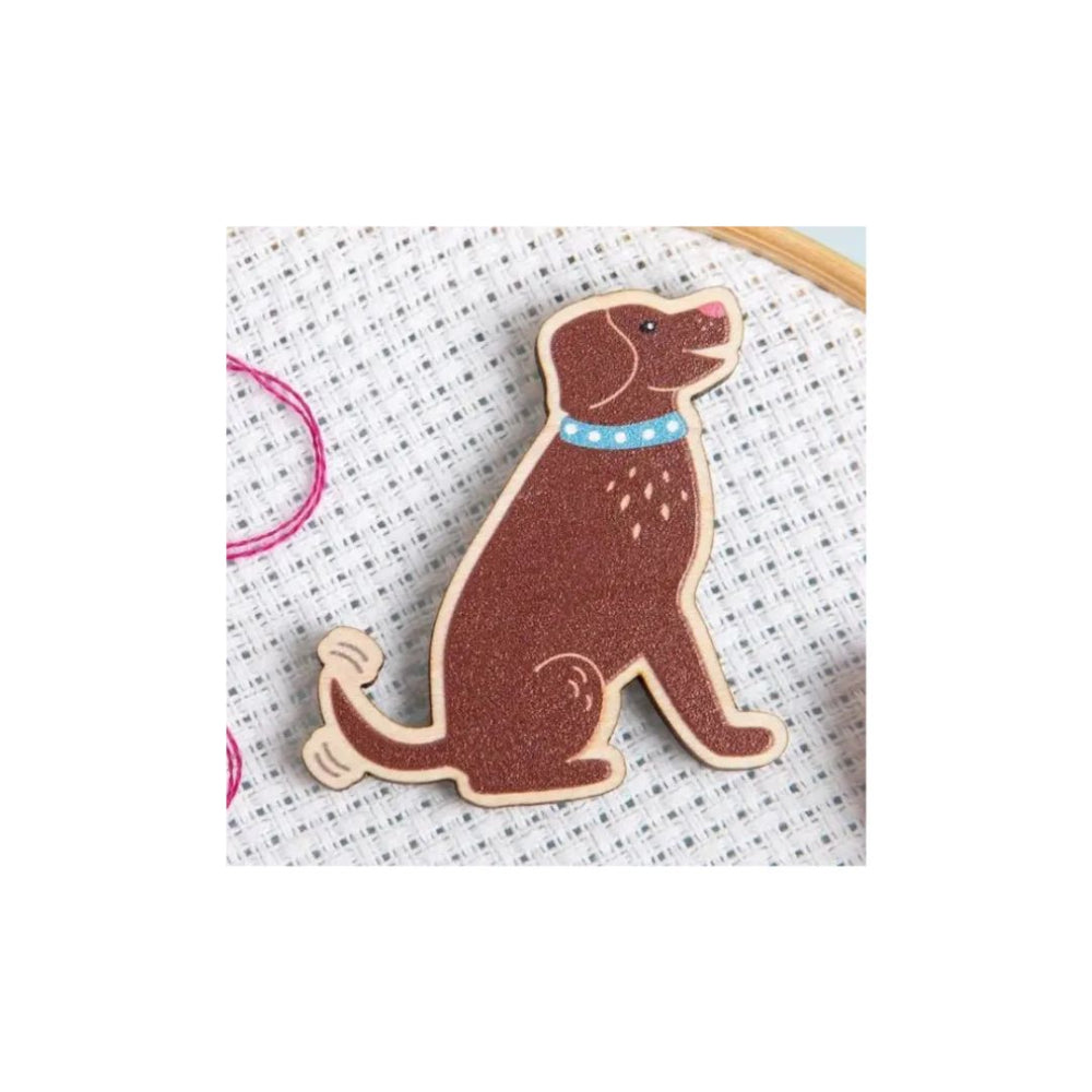 A brown dog-shaped wooden pin with a blue collar, displayed on a white cross-stitch canvas.