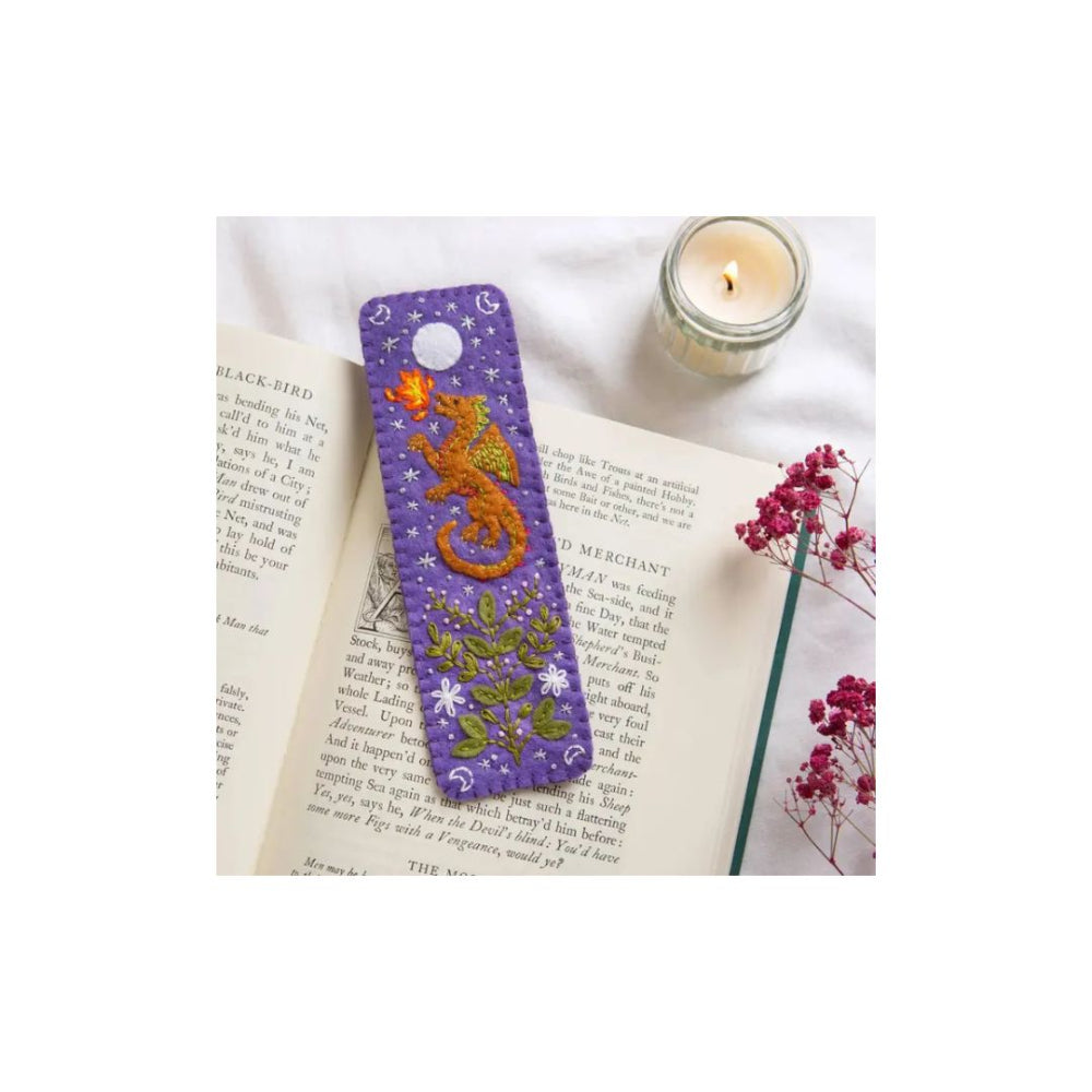 A purple book mark with a gold dragon and green foliage design, resting on an open book next to a lit candle and dried pink flowers.