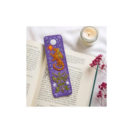A purple book mark with a gold dragon and green foliage design, resting on an open book next to a lit candle and dried pink flowers.