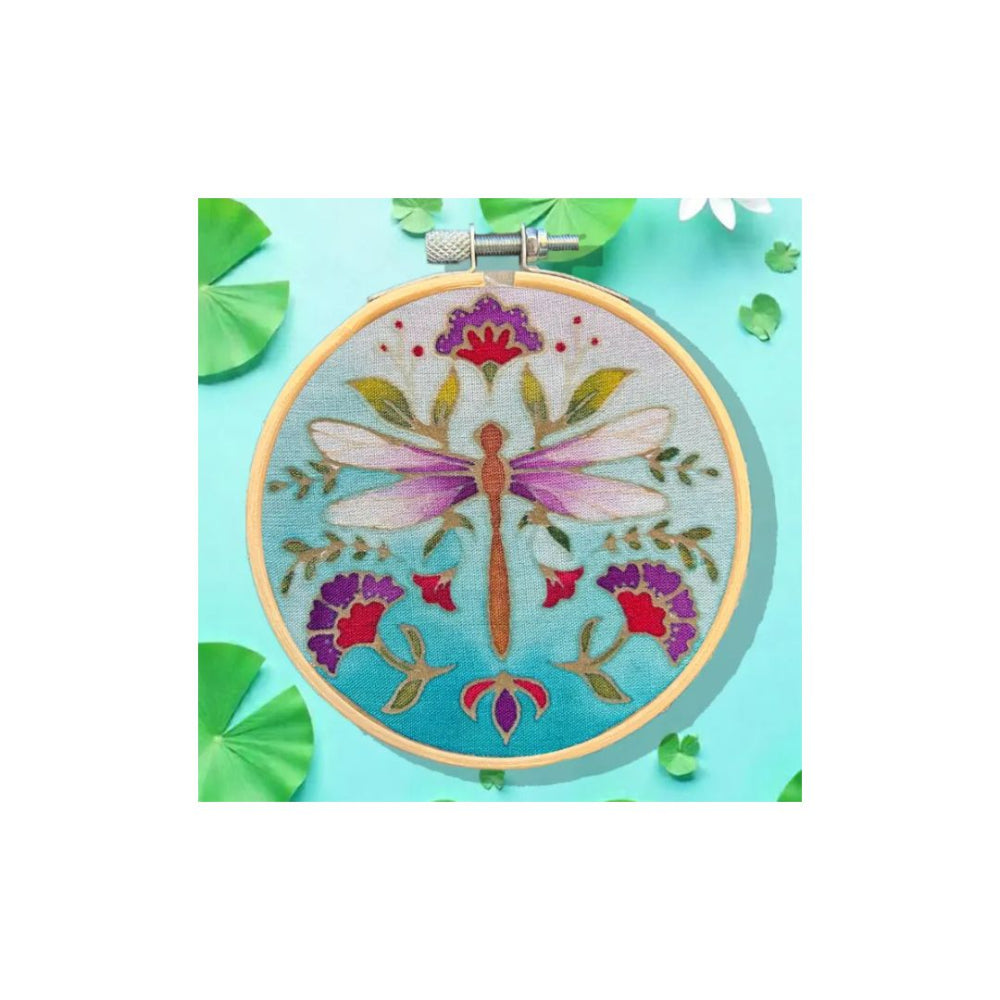 A circular embroidery hoop displaying a colourful dragonfly and floral design on light blue fabric, styled on a turquoise background with green leaf props.