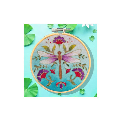 A circular embroidery hoop displaying a colourful dragonfly and floral design on light blue fabric, styled on a turquoise background with green leaf props.