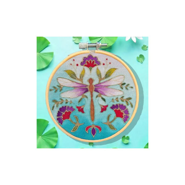 A circular embroidery hoop displaying a colourful dragonfly and floral design on light blue fabric, styled on a turquoise background with green leaf props.