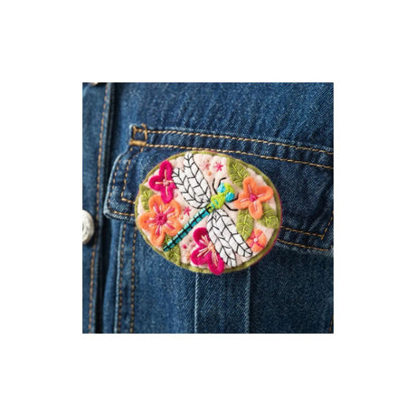 An oval embroidered patch featuring a white dragonfly with pink wings and colourful flowers, pinned onto a blue denim jacket pocket.