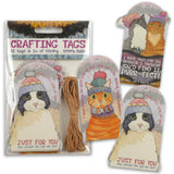 Pack of cat-themed crafting tags and string on a white background, with one tag shown unfolded and the others in the clear plastic package. The tags feature illustrated cats wearing knit hats and colorful text including 'CRAFTING TAGS' and 'JUST FOR YOU'.