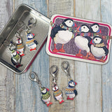 Three puffin-shaped crochet stitch markers with colourful knit-sweater designs are displayed on a rustic whitewashed wood surface, alongside a small metal tin and a matching decorative tin featuring puffin artwork.