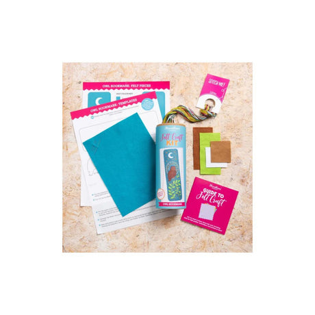 A flat-lay view of a DIY craft kit containing a light blue tin labeled 'The Craft Box', a teal sheet of paper, printed instruction sheets, a pink card, and small colored paper squares on a textured beige background.