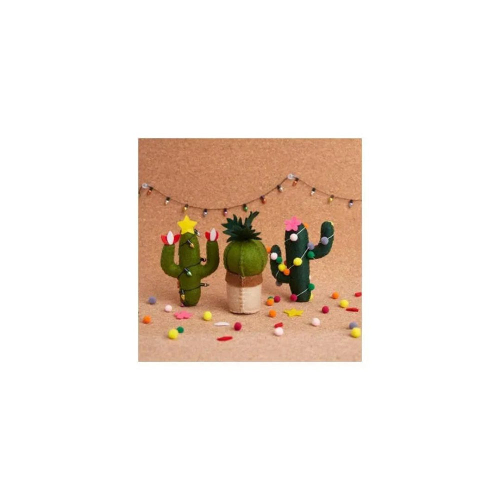 Three small felt cactus decorations in green and brown shades, decorated with colorful felt flowers and stars, arranged on a tan surface with small confetti and a string of fairy lights in the background.