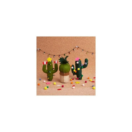 Three small felt cactus decorations in green and brown shades, decorated with colorful felt flowers and stars, arranged on a tan surface with small confetti and a string of fairy lights in the background.