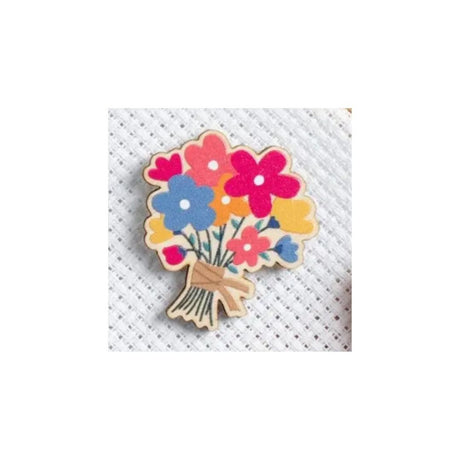 A colourful wooden pin shaped like a bouquet of flowers, featuring pink, blue, yellow, and red blossoms with a light brown wood-grain stem and bow, displayed on a white textured fabric background.