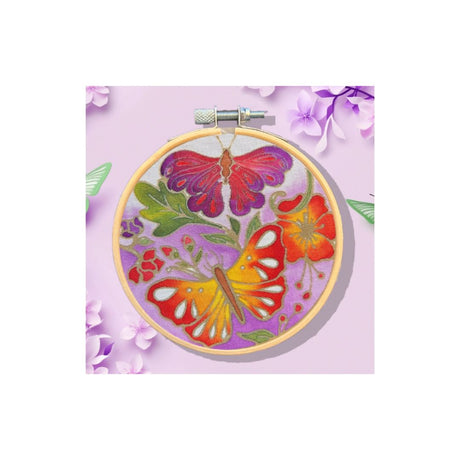 A circular embroidery hoop holding a colourful floral and butterfly design on lavender fabric, centered on a light lavender background with decorative purple flowers around the edges.