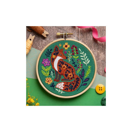 A round embroidery hoop containing a colorful stitched fox design on green fabric, photographed on a styled craft surface with spools of thread, ribbon, and buttons nearby.
